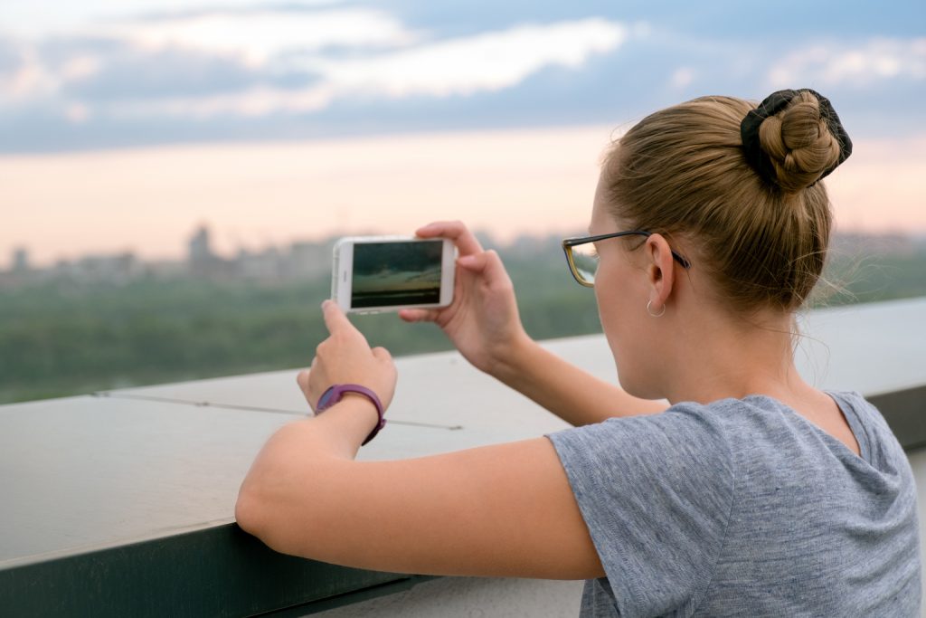 Girl blonde with glasses photographed on a white mobile phone on the roof of the house. Summer daytime