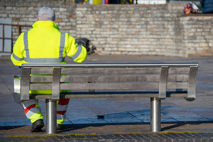 street cleaner wearing hi vis jacket and trousers has a quick coffee or tea break whilst sitting on a bench